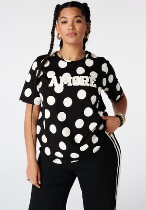 Femme avec deux tresses porte un t-shirt noir à gros pois blancs et le texte "AMORE", pantalon noir avec bandes blanches sur les côtés.