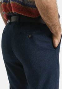 Dunkelblaue Hosen im Fischgrätmuster mit schmaler Passform, versehen mit einem Einzelknopfdetail und seitlichen Taschen. Brauer Gürtel sichtbar an der Taille.