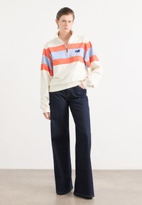 Kremfarget genser med oransje og lyseblå striper, halv-glidelås ved halsen, kombinert med mørkeblå vide jeans og minimale tilbehør.