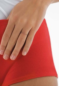 Anillo de oro delgado con pequeñas gemas redondas de color rosa engastadas en línea recta; mano descansando sobre una tela roja vibrante con una textura suave.