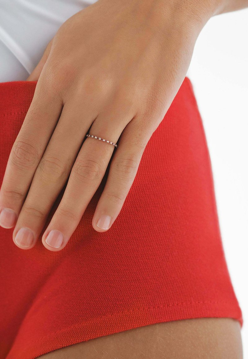 Anillo de oro delgado con pequeñas gemas redondas de color rosa engastadas en línea recta; mano descansando sobre una tela roja vibrante con una textura suave.