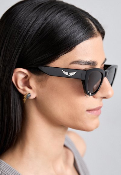 Lunettes de soleil noires angulaires avec des verres sombres, présentant un emblème en métal en forme d'aile sur la branche. Portées avec une boucle d'oreille décorative.