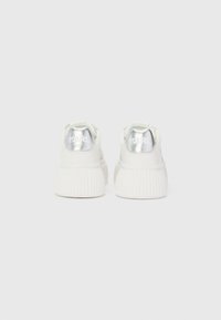 Baskets blanches avec une semelle texturée, présentant un accent argenté brillant sur le talon et une petite boucle pour un enfilage facile. Logo de la marque visible.
