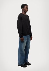 Črni pulover z širokim izrezom in dolgimi rokavi, v kombinaciji s širokimi modrimi jeans hlačami z blagim zbledenjem, obrabljenimi območji in črnimi čevlji.
