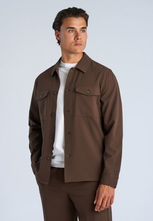 Mann trägt eine braune Jacke mit Knöpfen und passende Hose über einem weißen Hemd, steht mit den Händen in den Taschen und schaut zur Seite.