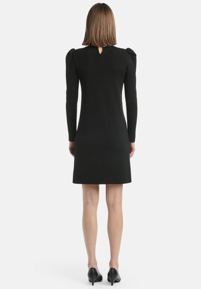 Schwarzes knielanges Kleid mit langen Ärmeln, strukturiertem Stoff, puffierten Schulterdetails und einem Rückenschlitz. Kombiniert mit schwarzen Absätzen.