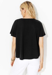 Femme aux cheveux blonds portant un haut noir à manches courtes ample et un pantalon blanc, de dos contre un fond blanc.