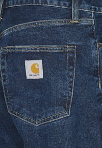 Jeans de mezclilla con un lavado azul oscuro. Las características incluyen costuras amarillas y un parche cuadrado blanco con un logo amarillo en el bolsillo trasero.