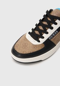 Pantofi sport cu model maro, cu accente negre și albe, având șireturile negre și o talpă din cauciuc. Logo-ul este afișat pe lateral. Textură netedă.