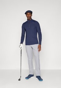 Golfantrekk med en marineblå kvart-zip topp, lysegrå bukser, hvit golvhanske, golfkølle og marineblå joggesko med blå akenter.