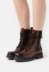 Bottines en cuir marron avec un motif texturé ressemblant à du crocodile, détail de fermeture éclair noire, bout rond et semelle en caoutchouc robuste.