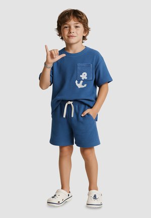 Young boy in blue shorts and t-shirt with anchor design, making a hand gesture, wearing white slip-on shoes with nautical prints.
