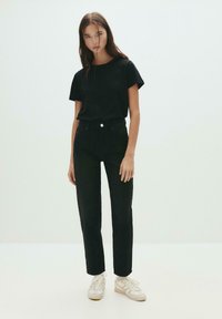 Schwarzes Baumwoll-T-Shirt kombiniert mit hochgeschnittenen schwarzen Jeans, beide figurbetont. Die Schuhe sind weiße und beige Sneaker mit strukturierten Akzenten.