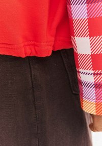 Czerwona bawełniana koszula z marszczonym dołem i kolorowym, kratkowanym długim rękawem z akcentami w kolorze fioletowym, czerwonym i pomarańczowym. Połączona z ciemnym dżinsem.