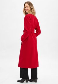 Donna che sorride sopra la spalla indossando un lungo cappotto rosso con cintura, pantaloni neri e stivali neri con tacco su uno sfondo bianco.