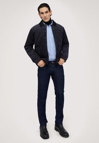 Chaqueta acolchada de color azul marino oscuro con un cuello negro, combinada con una camisa de rayas azul claro y jeans de mezclilla oscuros. Botas negras hasta el tobillo completan el conjunto.