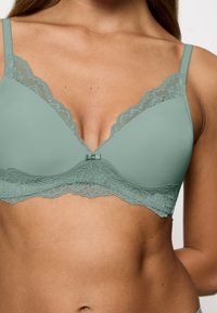 Bralette verde chiaro con pizzo, design a V, coppe morbide, un fiocco centrale e spalline regolabili. Tessuto liscio.