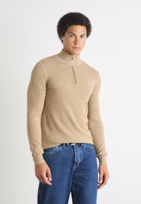 Maglione beige con zip in tessuto testurizzato, silhouette aderente e polsini a coste, abbinato a jeans di denim blu. Logo bianco sul petto.