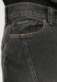 Κοντινή λήψη μαύρων τζιν παντελονιών που δείχνει την μπροστινή τσέπη, την τσέπη του νομίσματος, τον κρίκο ζώνης και τις λεπτομέρειες με πορτοκαλί ραφή.