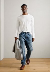 Hvit langermet skjorte, blå jeans, grå hettegenser drapert over armen, brune sko med tykke såler, står mot en lys vegg.