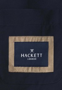 Tmavě námořnická látka se sešitým béžovým obdélníkovým záplatou a menší námořnickou záplatou zobrazující logo "Hackett London" s překříženými pálkami nad textem.
