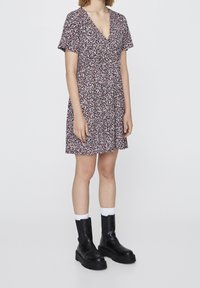 Robe fleurie en noir, rose et blanc, avec un décolleté en V, des manches courtes et une taille froncée. Portée avec des bottines noires épaisses et des chaussettes blanches.