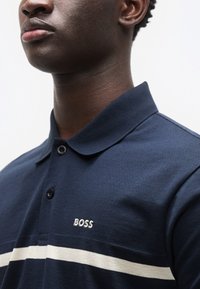 Μπλε ναυτικό πόλο πουκάμισο με υφασμάτινη υφή, γιακά με δύο κουμπιά και λευκή οριζόντια ρίγα με κεντημένο λογότυπο "BOSS" στο στήθος.