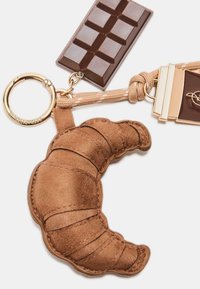 Un porte-clé en cuir marron représente un croissant, une barre de chocolat et un second accessoire, le tout attaché par un anneau en or.