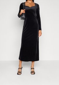 Robe maxi en velours noir avec des manches longues, un décolleté carré et une silhouette ajustée. Associée à des sandales à talons noires et un sac à main.