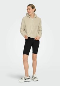 Beige fleecejakke med glidelås og hette, kombinert med svarte sykkelshorts og store hvite sneakers med svarte detaljer.
