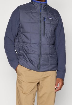 Mężczyzna ubrany w granatową pikowaną kamizelkę na długim rękawie oraz beżowe spodnie, stojący z jedną ręką w kieszeni na jednolitym tle.