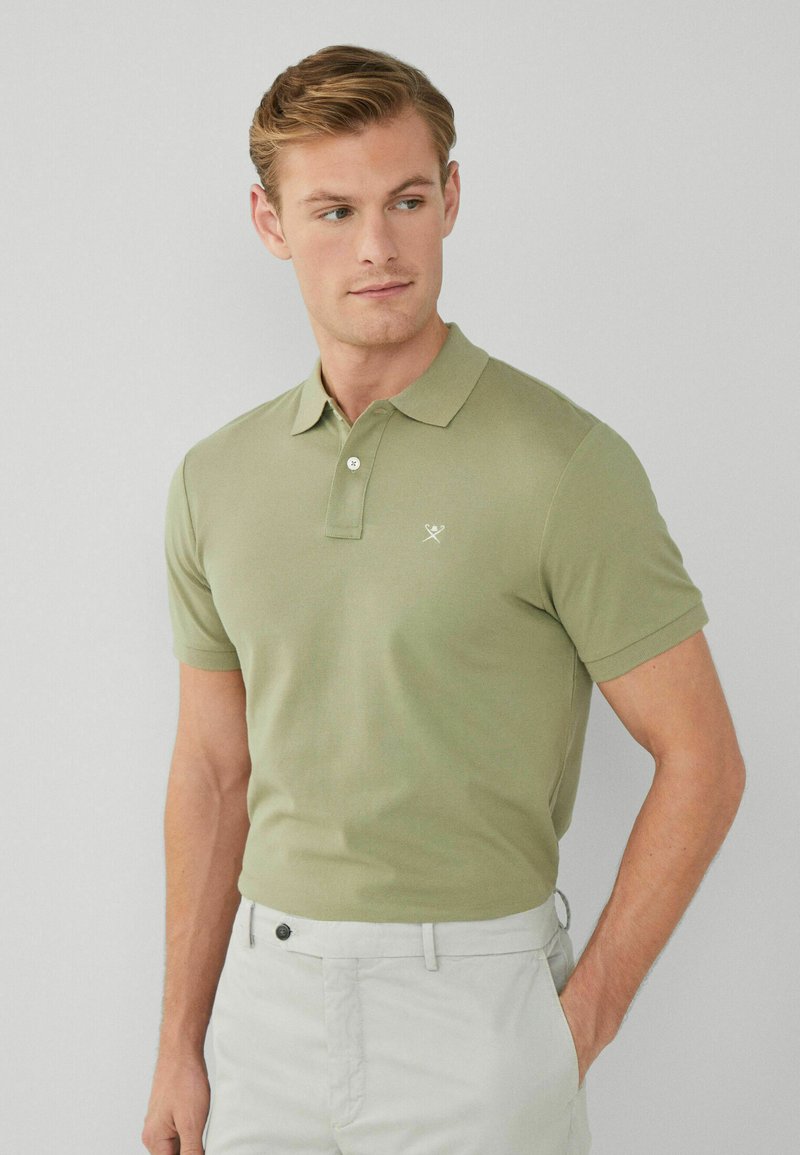 Olivgrön polo skjorta med korta ärmar, tvåknappsslutning och broderad logotyp. Modellen bär ljusa byxor.