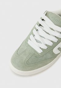 Baskets en suède vert avec des lacets blancs et une surface texturée, présentant un bout arrondi et des détails de couture subtils le long des côtés.