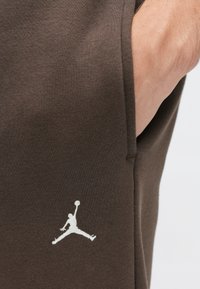 Brązowe bawełniane dżogery z boczną kieszenią, z białym haftowanym logo Jumpman blisko dołu. Gładka faktura, prosta sylwetka.