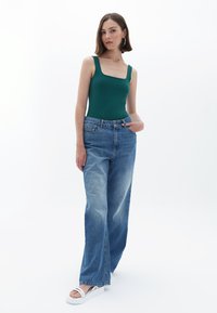 Body verde con scollatura a quadrato abbinato a jeans a gamba larga blu chiaro. Il modello indossa sandali con plataforma bianchi. Lo sfondo è bianco uniforme.