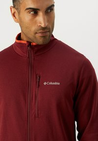 Hombre con una chaqueta Columbia de cremallera color burdeos con un pequeño bolsillo con cremallera en el pecho, mirando hacia un lado contra un fondo liso.