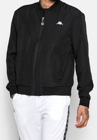 Giubbotto bomber nero realizzato in tessuto testurizzato, dotato di chiusura con zip, polsini a costine, due tasche laterali e un logo bianco sul petto.