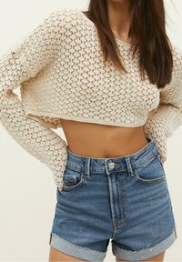 Suéter corto de punto de crochet, color crema, con mangas largas, combinado con pantalones cortos de mezclilla azul claro de talle alto con dobladillos enrollados.