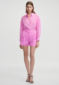 Camisa de lino rosa con botones y mangas largas acompañada de pantalones cortos de talle alto a juego. El modelo lleva sandalias negras con tiras en los tobillos.