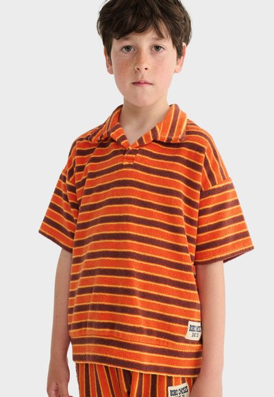 Niño pequeño con camisa de manga corta a rayas naranja y marrón y pantalones cortos a juego, de pie frente a un fondo liso.