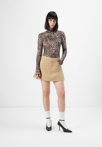 Vrouw met steil zwart haar, gekleed in een langwerpige luipaardprint top, een beige rok, witte sokken en zwarte hakken, staande tegen een witte achtergrond.