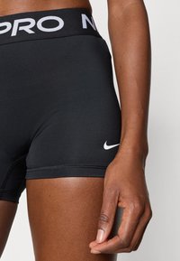 Μαύρα αθλητικά σόρτς από ελαστικό υλικό, με λευκό λογότυπο και ζώνη μέσης με τη μάρκα. Η κοντινή λήψη δείχνει τη λεπτομέρεια της ραφής.