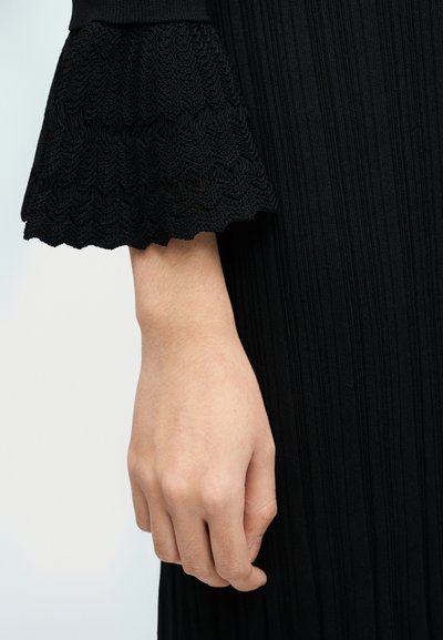 Αριστερό χέρι χαλαρό στο πλάι, φοράει μαύρο ριμπ φόρεμα με φαρδύ μανίκι με υφή που τελειώνει πάνω από τον καρπό.