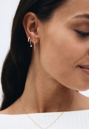 Donna che indossa tre orecchini d'oro all'orecchio: uno con piccoli diamanti, uno con una perla e uno con un pendente a spiga.