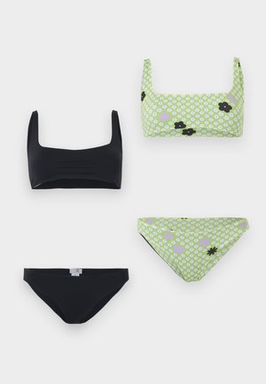Kvinne står barfot og har på seg en lys grønn blomstret bikini med hvite og svarte blomster mønstre mot en ensfarget hvit bakgrunn.