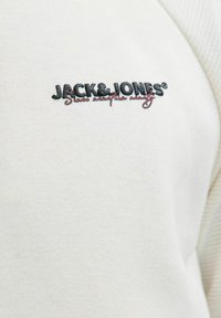 Bela dukserica s pleteno teksturo, ki ima na prsih izvezen logo "JACK & JONES" v črni in rdeči barvi, ki prikazuje preproste oblikovalske detajle.