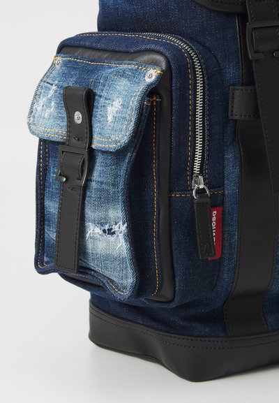 Geantă din denim cu accente din piele neagră, având un buzunar frontal din denim uzat, accesorii din metal argintiu și cusături galbene în contrast.