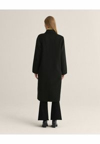 Lang sort trenchcoat med høj krave, lige pasform og sænkede skuldre, parret med flagrende sorte bukser og sorte støvler.