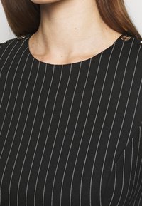 Černý textilní top s vertikálními bílými pruhy, kulatým výstřihem a malými zlatými knoflíky na ramenních akcentovacích detailech. Hladká textura.