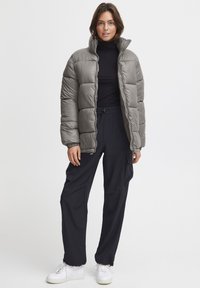 Grå puffjacka med hög krage, kviltad design och dragkedjestängning. Matcher med svarta byxor och vita sneakers, vilket skapar en lager-på-lager-look.